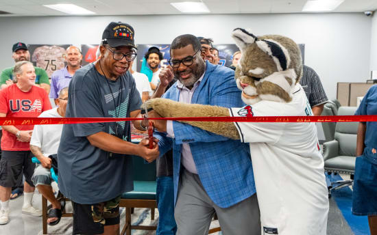 A person in a blue plaid blazer and another in a dark t-shirt and camo shorts cut a red ribbon with large scissors, assisted by a mascot in a white sports jersey, indicating a ceremonial event.