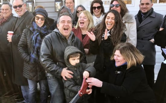 Group of people outdoors dressed in winter coats and scarves, gathered around a child holding large red-handled scissors, suggesting a ribbon-cutting or ceremonial event.