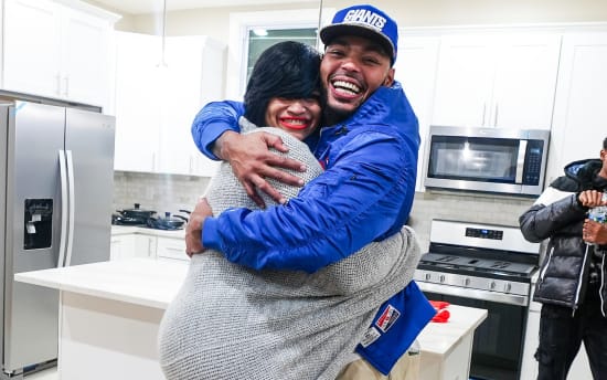 Two people embracing in a modern white kitchen with stainless steel appliances, including a refrigerator, microwave, and stove. One person wears a blue jacket and a blue Giants cap, while the other wears a gray knit sweater. Another person in a black and white jacket holds a water bottle in the background.