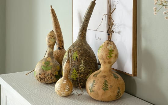 Decorative dried gourds with natural beige and brown tones, featuring hand-painted green leaves and white flowers, displayed on a light wood surface.