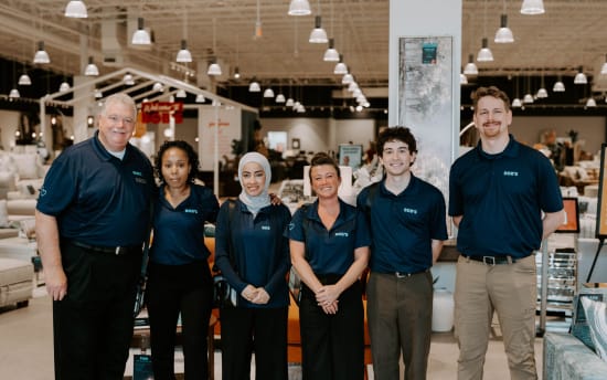 Six retail employees wearing navy blue polo shirts with "BOB'S" logo stand inside a well-lit furniture store with sofas and decor in the background.