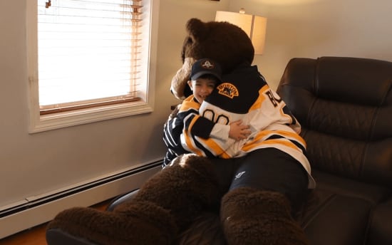 A person hugging a large brown bear mascot wearing a Boston Bruins hockey jersey, sitting on a black leather chair near a window with blinds.