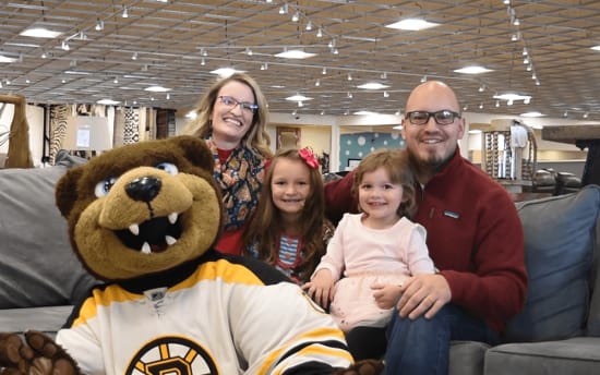 A person in a Boston Bruins mascot costume with a large brown bear head and white hockey jersey sits on a gray couch with three children and an adult wearing a red sweater in a spacious, well-lit indoor setting.