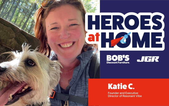 Woman with a light-colored dog outdoors next to a "Heroes at Home" banner featuring sponsors Bob's Discount Furniture and JGR, highlighting Katie C., Founder and Executive Director of Resonant Vibe.