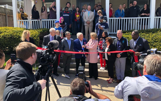A ribbon-cutting ceremony in front of a house with a group of people, including officials and media, capturing the event.