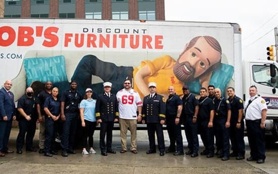 Group of uniformed emergency responders and staff standing in front of a large white truck with "BOB'S DISCOUNT FURNITURE" and "MYBOBS.COM" branding, featuring an advertisement of a person reclining on a blue chair.
