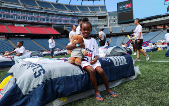 Child sitting on a New England Patriots-themed sleeping bag on a football field, hugging a teddy bear wearing a Patriots sweatshirt, with other children and adults in the background.