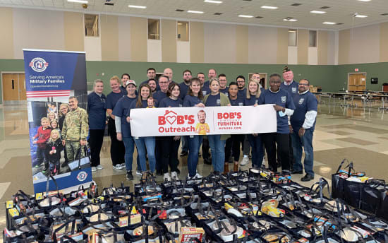 Group of people wearing navy blue shirts holding a banner that reads "BOB'S Outreach BOB'S DISCOUNT FURNITURE MYBOBS.COM" standing behind numerous black reusable bags filled with food and supplies, next to a vertical banner that says "Serving America's Military Families OperationHomefront.org" in a large indoor space.