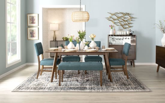 Dining room setup with mid-century modern wooden table and blue upholstered chairs and bench, decorative vases with flowers, patterned area rug, and wall art.