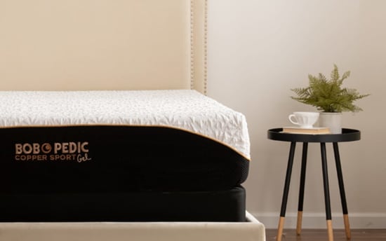 Mattress with beige upholstered headboard and black base, featuring "BOBOPEDIC Copper Sport Gel" branding on the side, next to a black round side table with wooden legs, a white cup, a book, and a green potted plant.