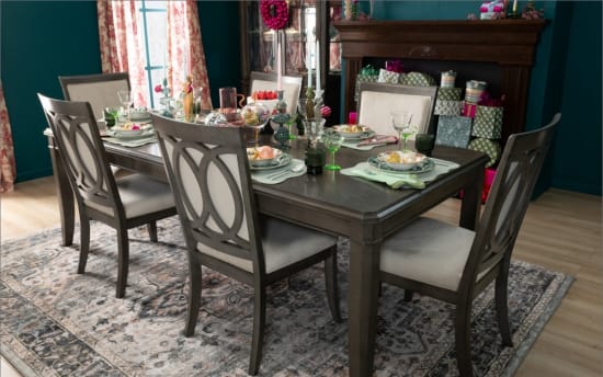 Dark wood dining table set with four cushioned chairs on a patterned rug, decorated with glassware, plates, and holiday ornaments.
