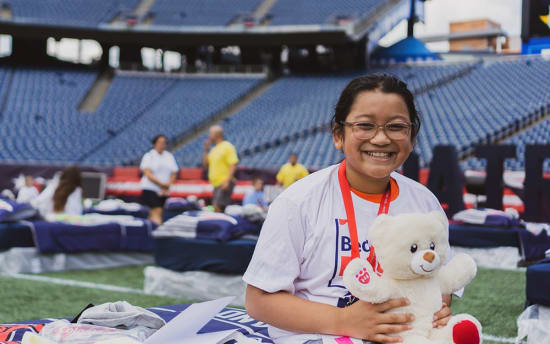A person wearing a white t-shirt with a red lanyard sits in a stadium holding a white plush teddy bear with red paw prints and feet.