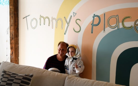 Man and child in astronaut costume giving thumbs up, standing in front of a colorful wall mural with "Tommy's place" text.