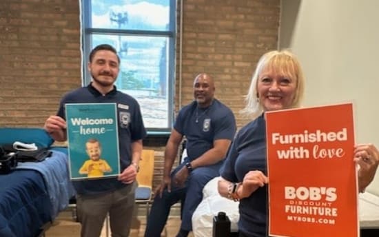 Three people in a dorm room holding signs; one reads "Welcome home" with a cartoon character, the other "Furnished with love BOB'S DISCOUNT FURNITURE MYBOBS.COM," promoting furnished living spaces and Bob's Discount Furniture.