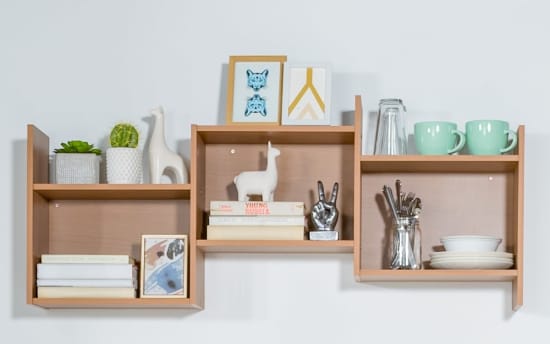 Wooden wall-mounted shelving unit with three compartments holding decorative items: potted succulents, ceramic llama figurines, framed art, books, metal peace hand sculpture, glassware, mint green mugs, and white dishware.
