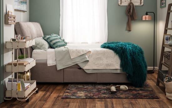 Modern bedroom with a gray upholstered bed, teal textured throw blanket, patterned area rug, wooden floor, and soft green walls.