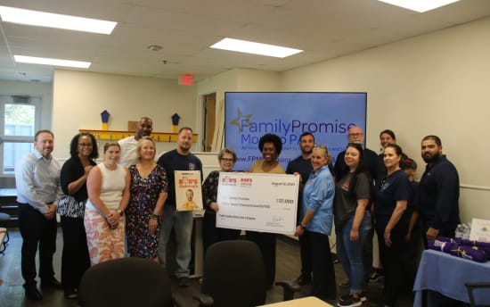 A group of people standing indoors holding a large check for $37,000 dated August 12, 2024, from Bob's Outreach to Family Promise, with a TV screen in the background displaying "Family Promise" and related text.