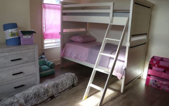 White wooden bunk bed with pink and purple bedding in a child's bedroom, featuring a ladder, dresser, and Minnie Mouse-themed storage organizer.