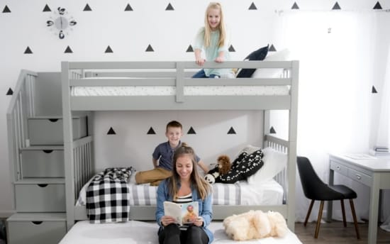 Modern gray wooden bunk bed with stairs, featuring striped and checkered bedding, a child reading a book, and a fluffy white dog on a pull-out trundle bed in a bright, minimalist bedroom.