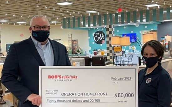 Two people holding a large check from Bob's Furniture dated February 2022, payable to Operation Homefront for $80,000, with the amount written as "Eighty thousand dollars and 00/100." The background shows a café area with tables and chairs.