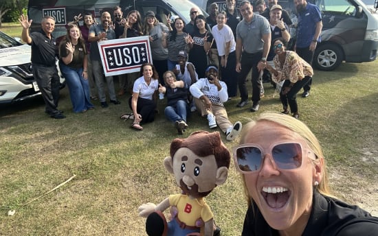Group of people outdoors holding a USO sign near vans with USO logos, with one person holding a plush doll with brown hair, beard, yellow shirt, and blue pants.