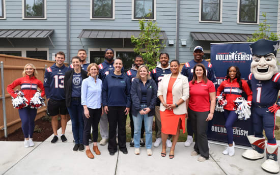 Group of people posing outdoors in front of a gray building with windows, including individuals in New England Patriots jerseys, cheerleader outfits, and casual attire, standing near a banner that reads "Celebrate Volunteerism Patriots Foundation."