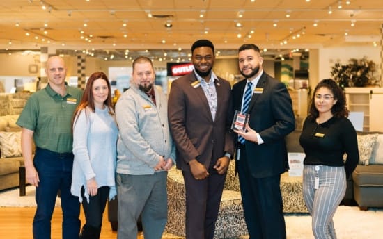 Six retail employees standing in a furniture store showroom, dressed in business casual and formal attire, holding an award plaque.