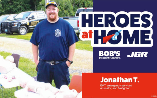 Man in navy blue firefighter uniform stands outdoors with rescue training dummies on tables. Text reads: "HEROES at HOME, BOB'S Discount Furniture, JGR, Jonathan T. EMT, emergency services educator, and firefighter." Sponsored by Joe Gibbs Racing.