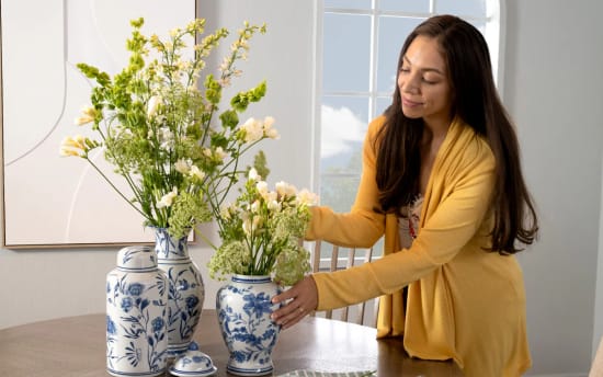 Woman arranging fresh green and white flowers in blue and white ceramic vases on a wooden dining table near a window, natural light.