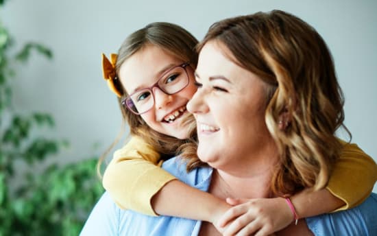 A child in a yellow long-sleeve shirt hugging an adult wearing a light blue shirt, showing a warm family moment indoors with a blurred green plant background.