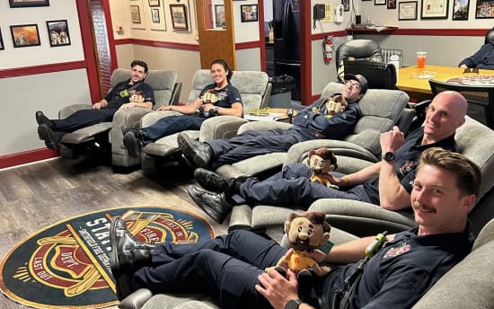 Firefighters relaxing on gray recliner chairs inside a fire station lounge with plush firefighter mascot toys, wood flooring, and framed certificates on walls.