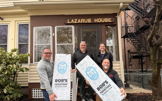 Four people holding large white boxes with blue graphics and text "BOB'S DISCOUNT FURNITURE MYBOBS.COM" stand in front of a building with a sign "LAZARUS HOUSE" above the door, indicating a furniture donation or delivery.
