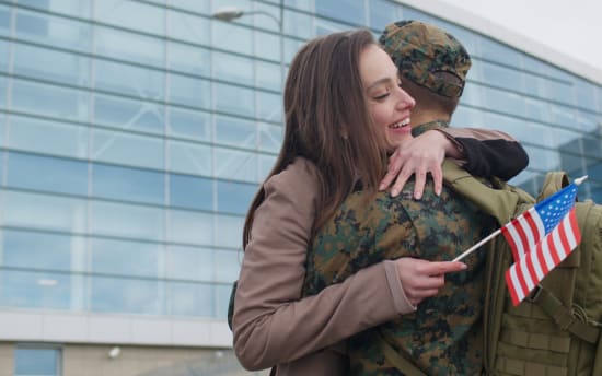 A woman warmly hugging a soldier in camouflage uniform holding a small American flag, outside a modern glass building.