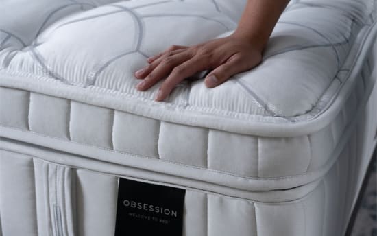 Hand pressing a white, quilted mattress topper with a visible label reading "OBSESSION WELCOME TO BED," highlighting mattress comfort and brand.