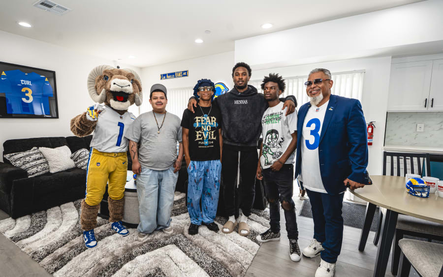 Group of six people standing indoors on a patterned rug, including a person in a Los Angeles Rams mascot costume, casual clothing with graphic tees, and one in a blue blazer with a white jersey featuring a large blue number 3. A framed blue Rams jersey with "CURL 3" hangs on the wall. The setting appears to be a casual fan or team event in a bright, modern room.
