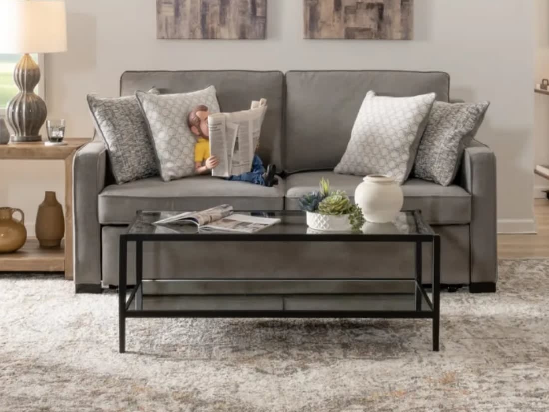 Gray fabric loveseat sofa with four patterned throw pillows, a glass coffee table with decorative plants and a vase, and a side table with a lamp in a modern living room.