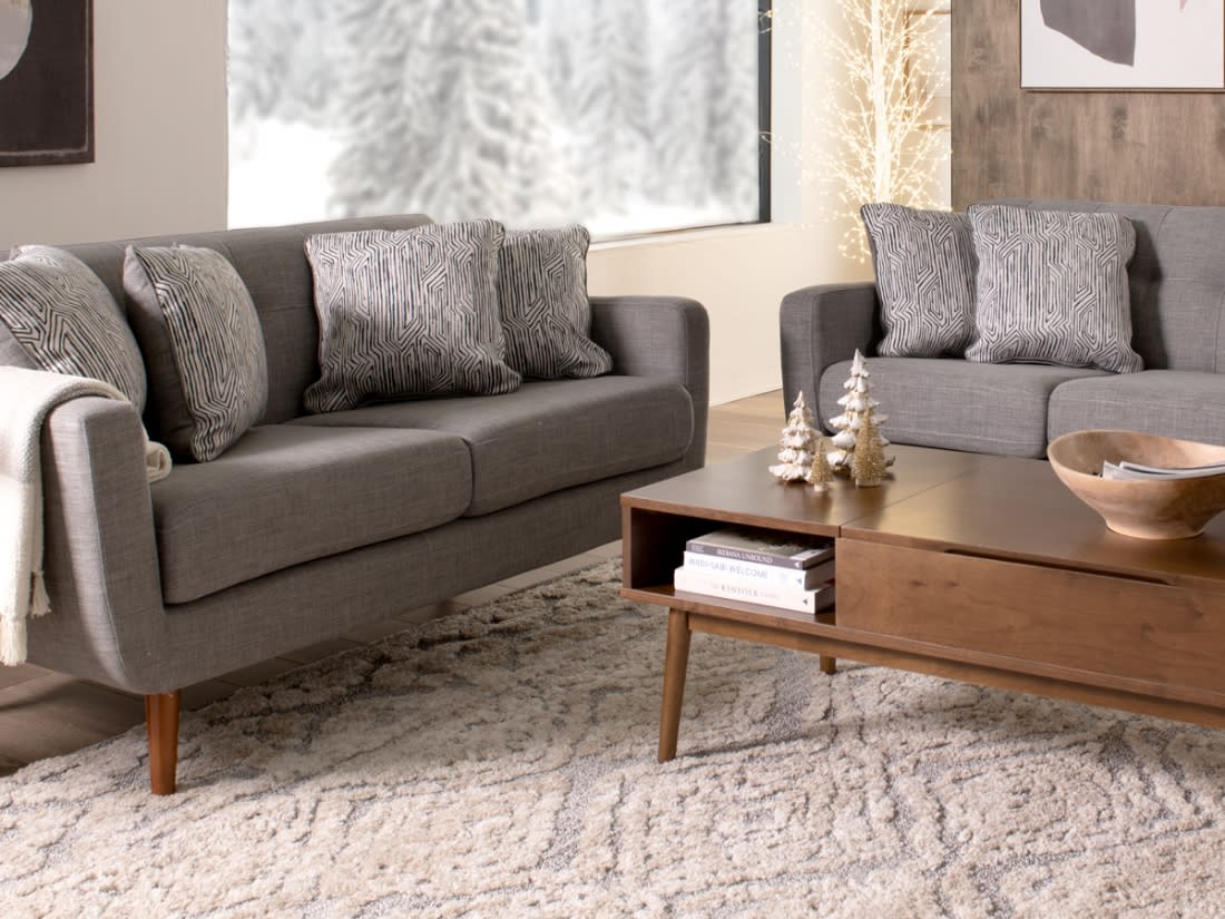 Modern living room with two gray fabric sofas featuring patterned cushions, a wooden mid-century coffee table with decorative trees, and a textured beige rug.