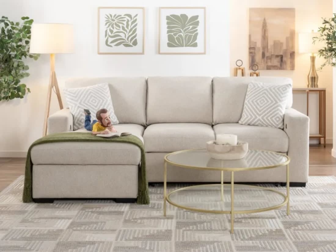 Modern living room setup with a beige fabric sectional sofa, matching ottoman with green throw, glass coffee table with gold frame, and decorative plants and lamps.