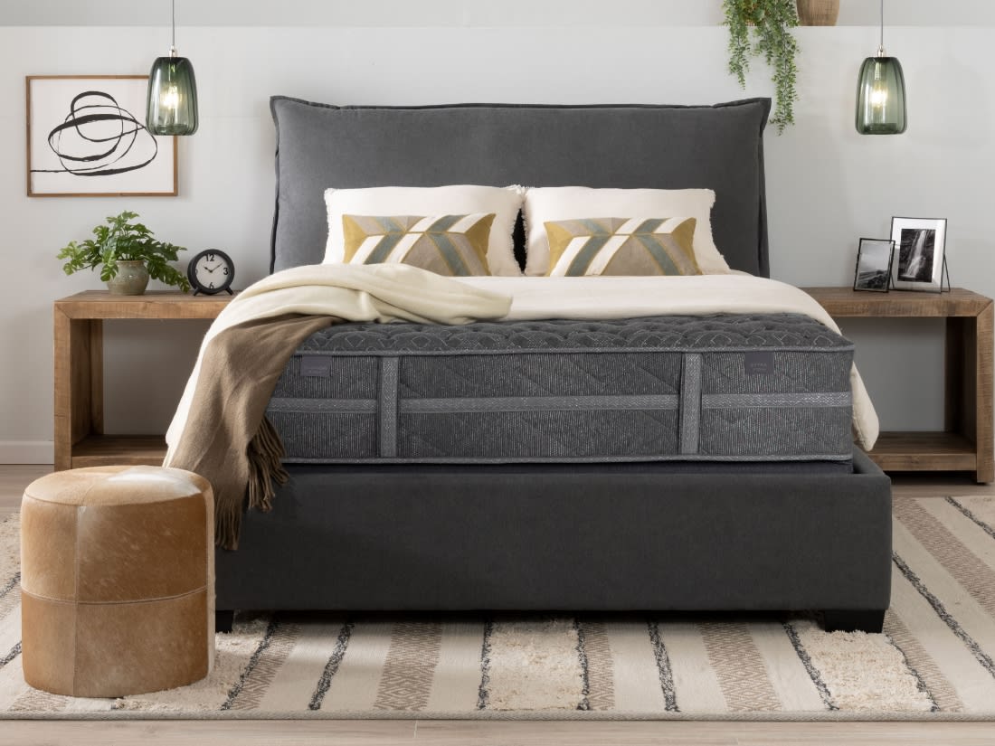 Gray upholstered bed with a plush headboard, thick gray mattress, cream and gold geometric pillows, beige throw blanket, wooden nightstands, and a tan leather ottoman on a striped rug.