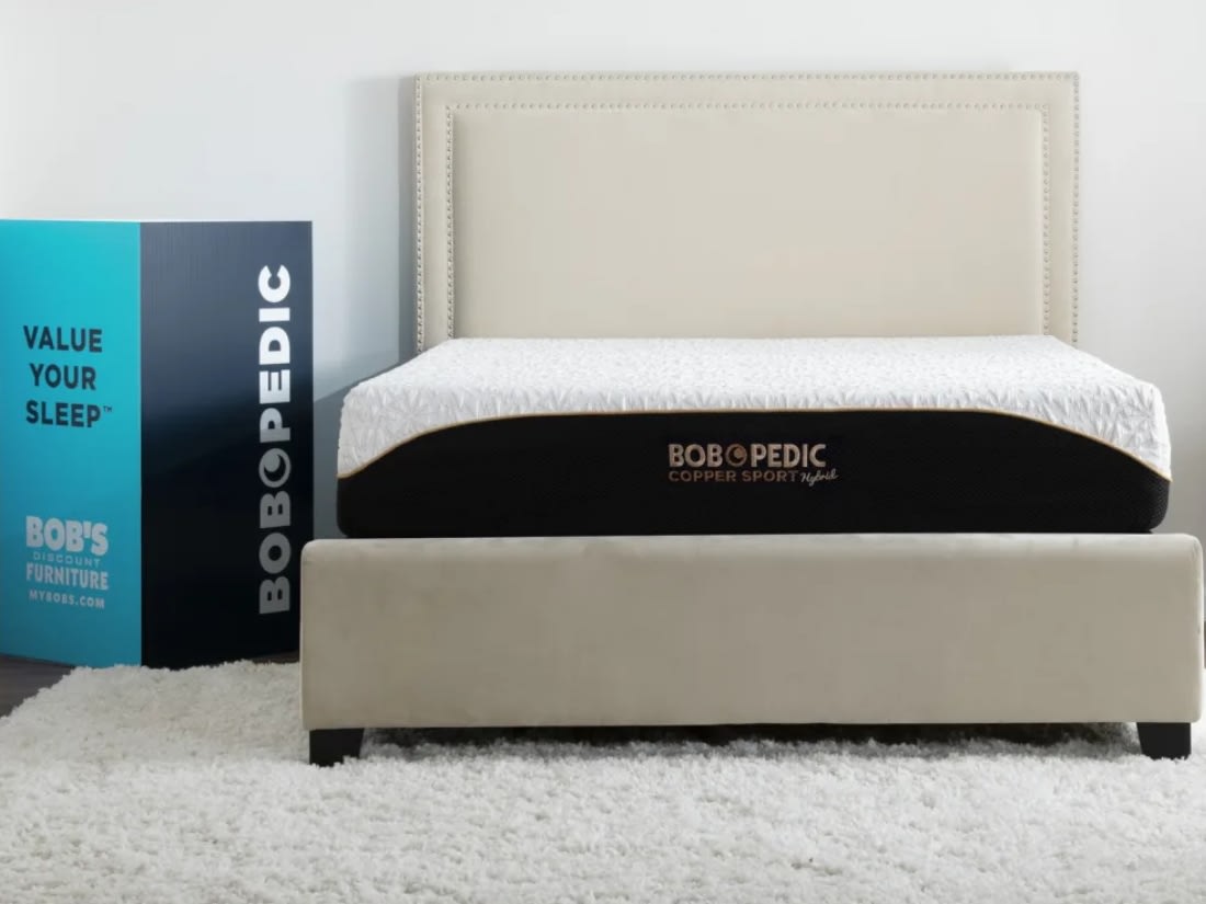 BOBOPEDIC Copper Sport Hybrid mattress on beige upholstered bed frame with nailhead trim, next to blue box with text "VALUE YOUR SLEEP™ BOBOPEDIC BOB'S DISCOUNT FURNITURE MYBOBS.COM"