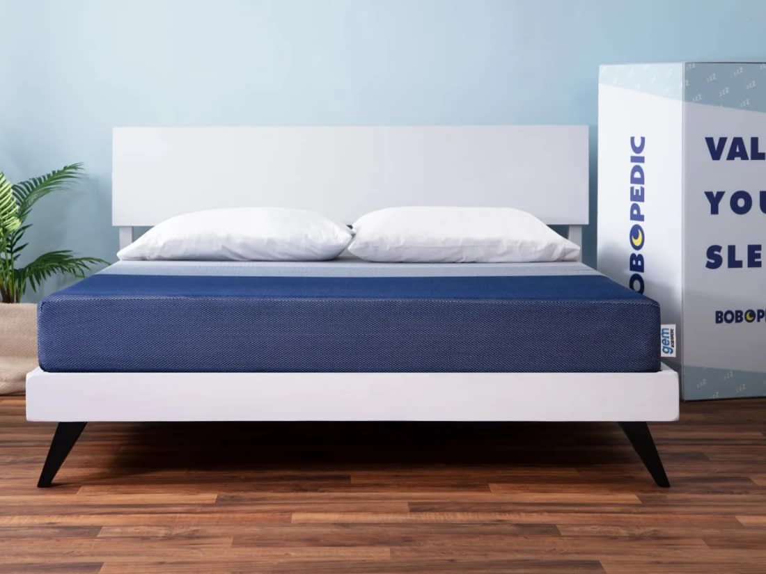 Modern bed frame with white wooden headboard and base, navy blue memory foam mattress labeled "gem," two white pillows, wooden floor, and blue wall. Box on right side reads "BOBOPEDIC" vertically and "VALUE YOUR SLEEP" horizontally, promoting Bobopedic mattress brand.