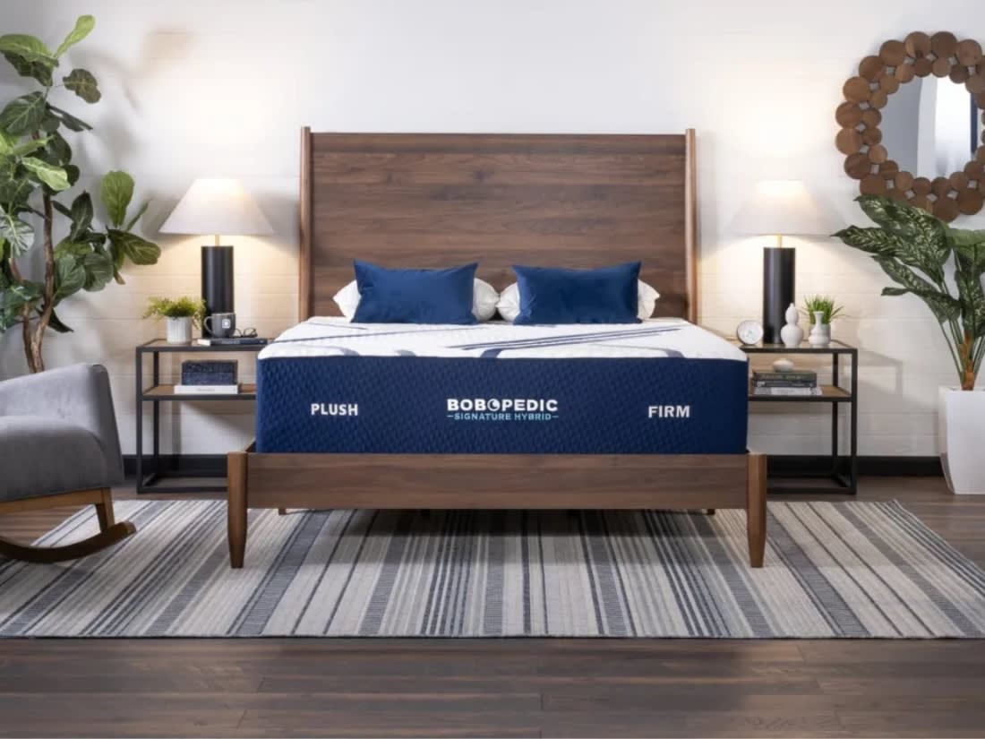 King-size wooden bed frame with a BOBOPEDIC Signature Hybrid mattress labeled "PLUSH" and "FIRM," featuring blue pillows, modern nightstands, lamps, and decorative plants.