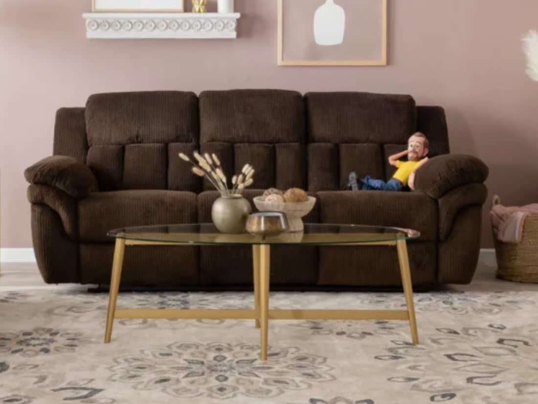 Dark brown plush fabric reclining sofa with three seats, thick padded armrests, and a glass coffee table with wooden legs in a modern living room setting.