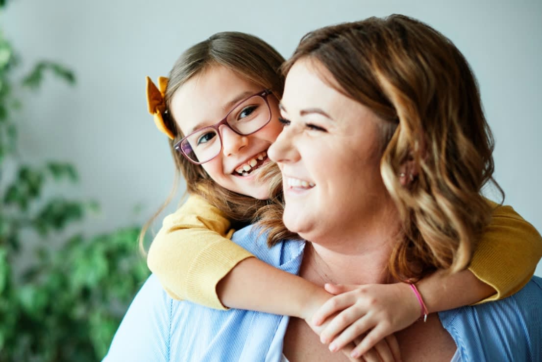 A child in a yellow long-sleeve shirt hugging an adult wearing a light blue shirt, showing a warm family moment indoors with a blurred green plant background.