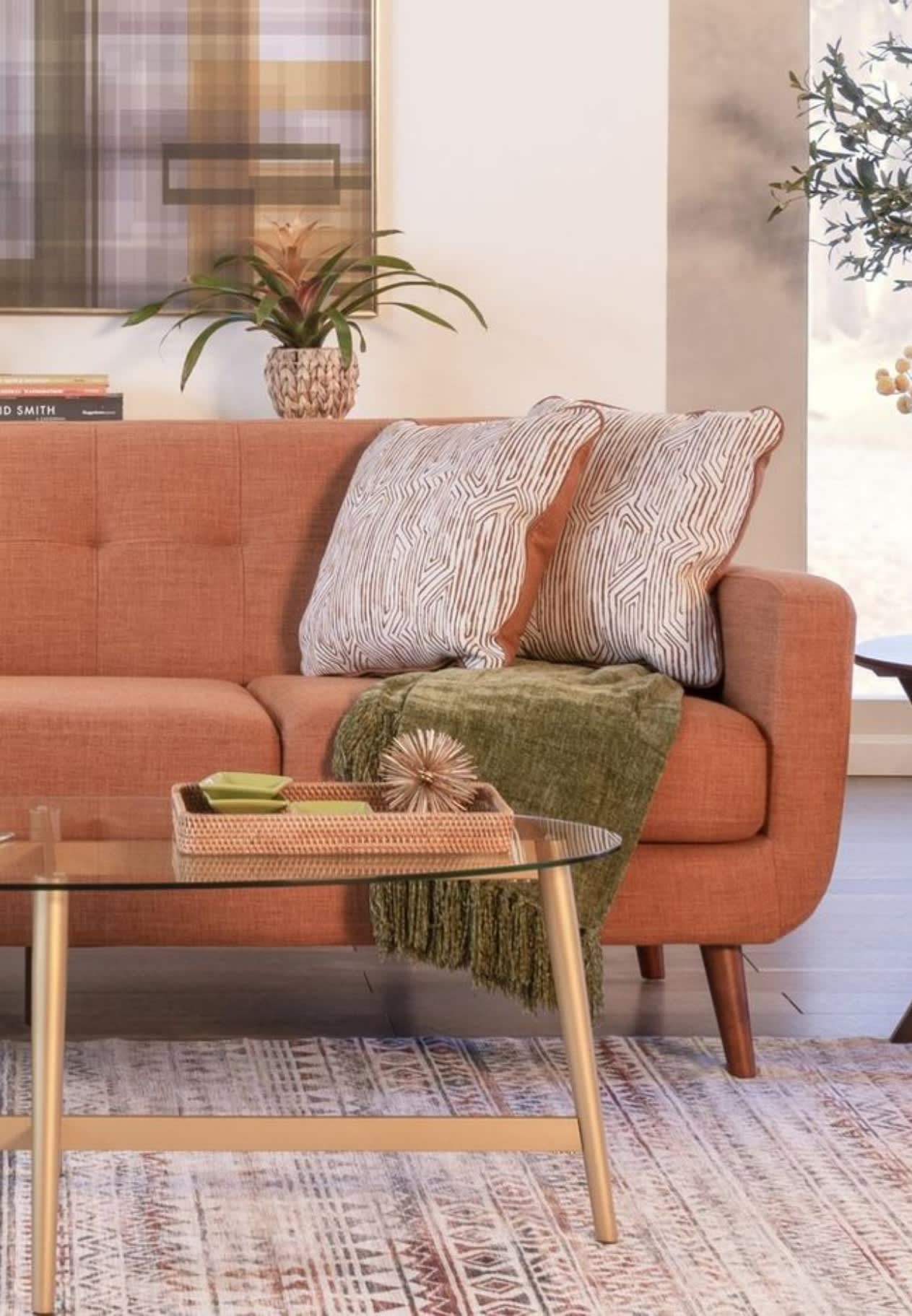 Rust-colored fabric sofa with two patterned beige and brown throw pillows, an olive green textured throw blanket, glass coffee table with gold legs, and a decorative tray.
