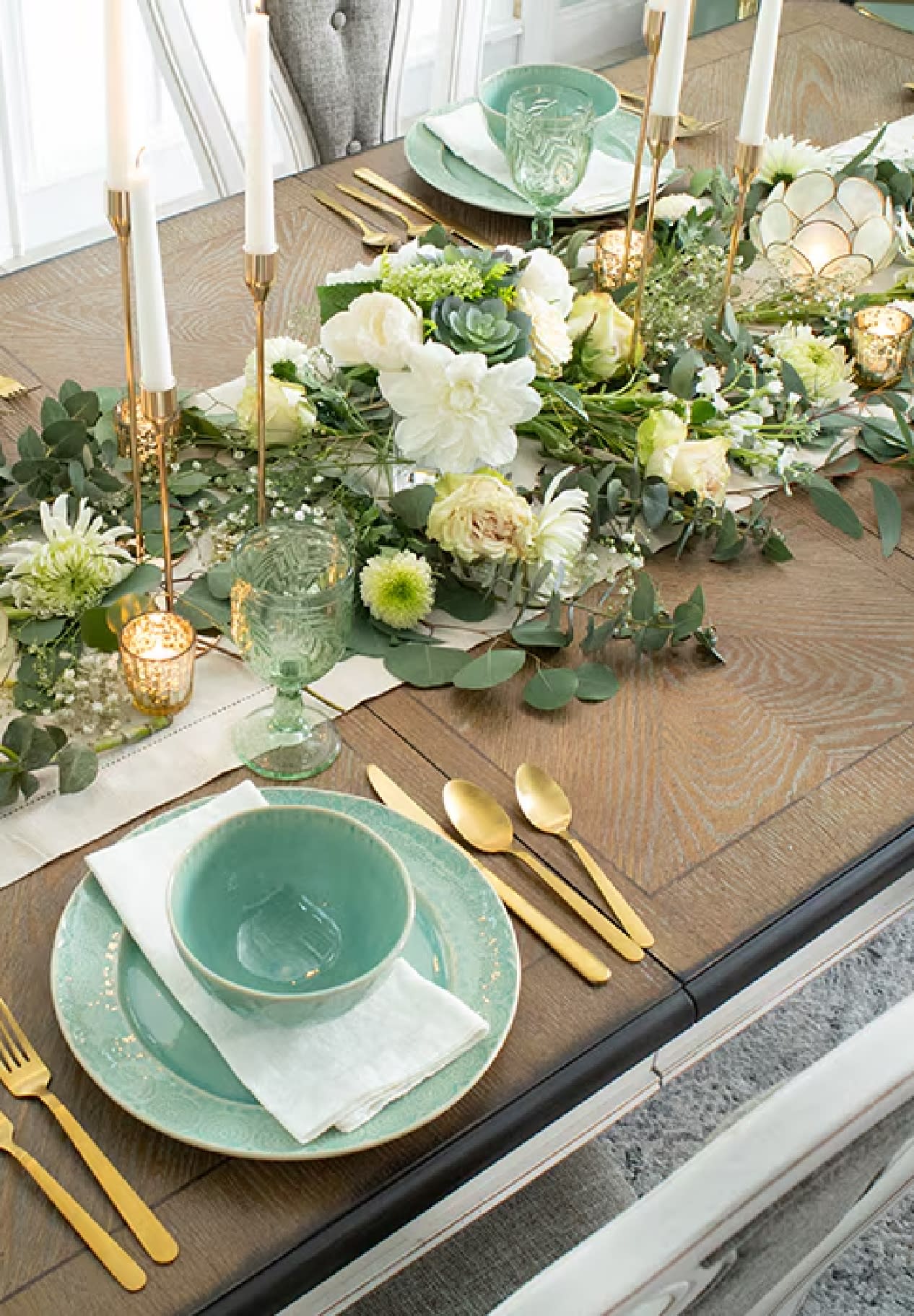 Spring inspired tablescape with flowers arranged on a table runner, white taper candles in gold candle holders, and mint colored dinnerware with gold cutlery