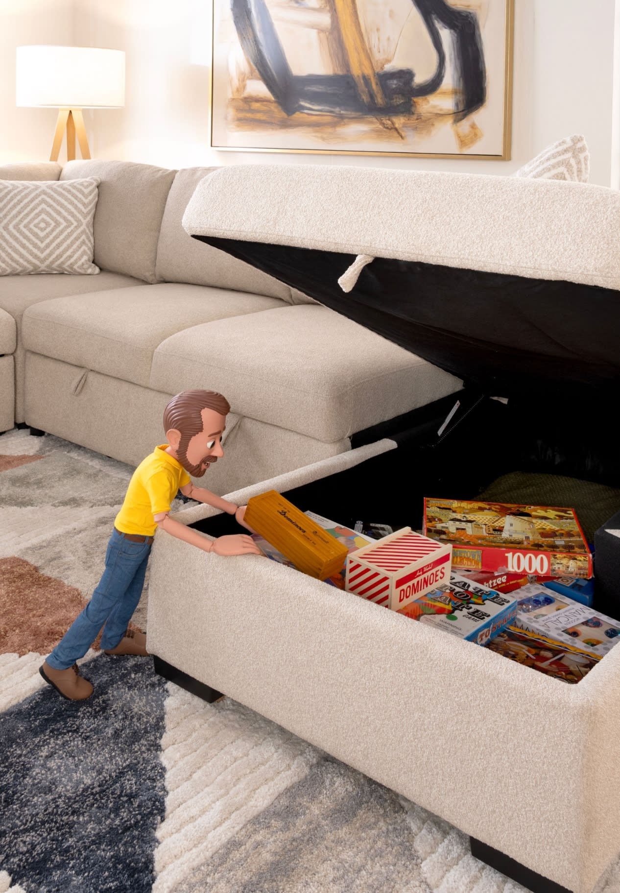 Beige fabric storage ottoman with lift-up lid revealing board games inside, including Dominoes and a 1000-piece puzzle, in a modern living room.