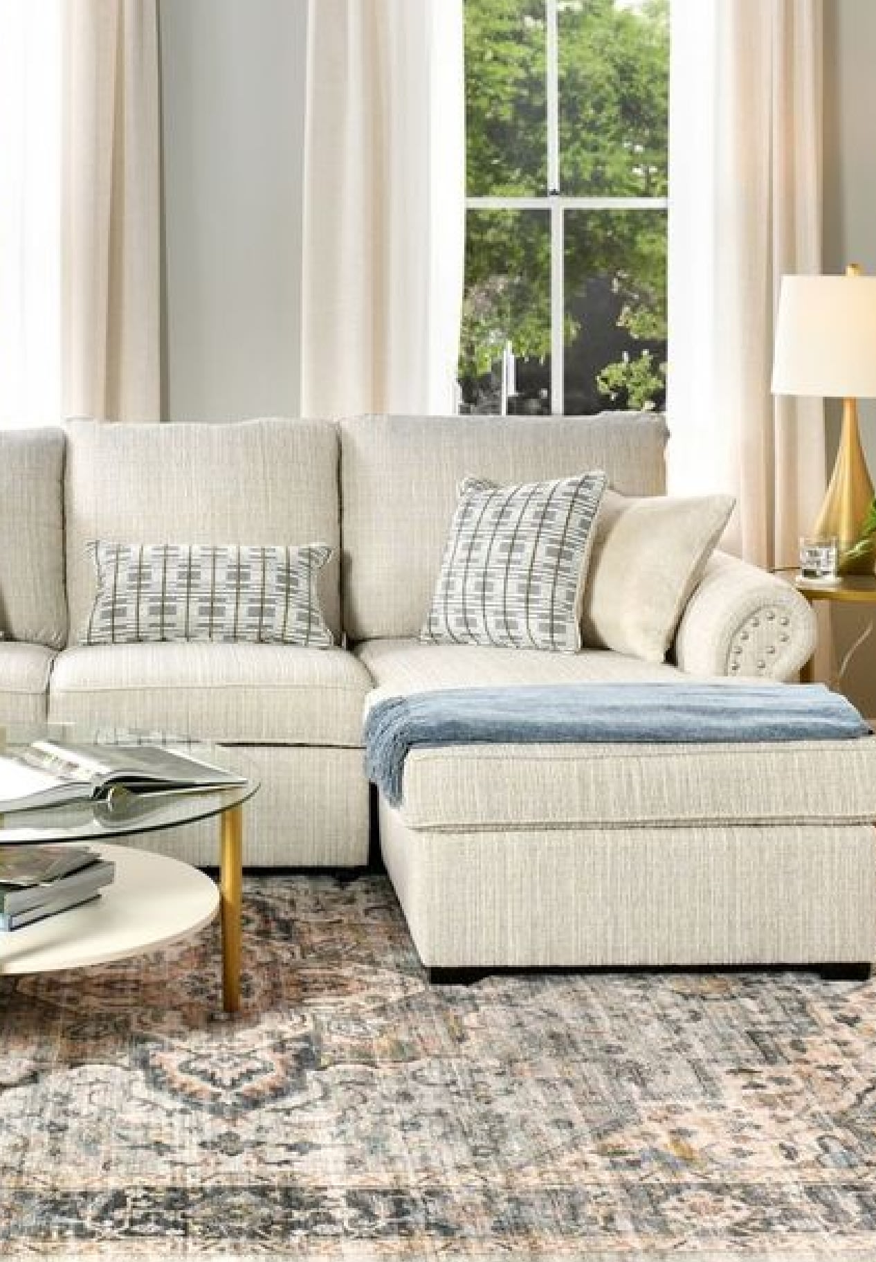 Beige sectional sofa with textured fabric, patterned throw pillows, blue throw blanket, glass and white round coffee table, and a patterned area rug in a bright living room.