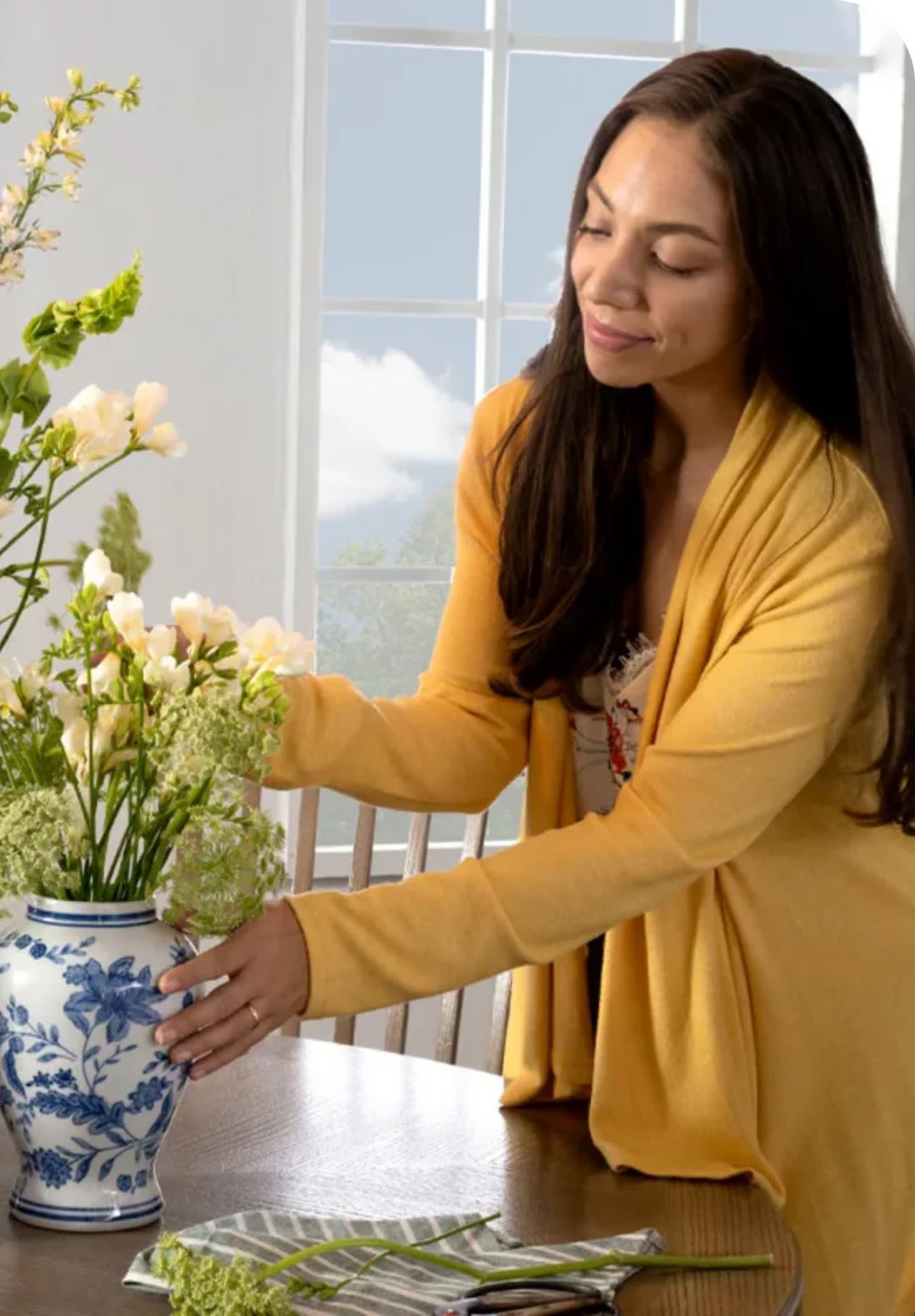 Woman arranging white and green flowers in a blue and white ceramic vase on a wooden table near a window, wearing a mustard yellow cardigan.