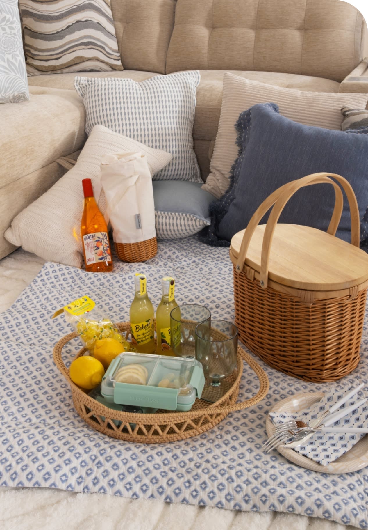 Cozy beige sectional sofa with assorted textured pillows, blue patterned picnic blanket, wicker tray with lemons, Belvoir lemonade bottles, glasses, and a picnic basket.